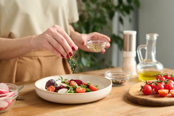 A cook seasoning with spices from a sauce bowl the salad in a white dish standing on the table next to cherry tomatoes on a cutting board and a jug of vegetable oil