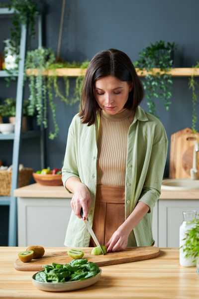 A woman with short dark hair in a light outfit cuts kiwi for making smoothie on a wooden cutting board next to which spinach is in an oval dish