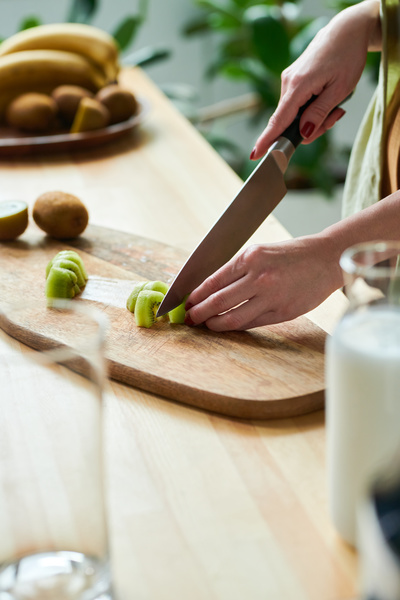 A female smoothie maker slices peeled kiwi for making smoothie with a large knife on a cutting board next to which the ingredients stand