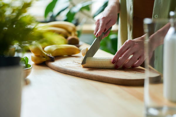 A female smoothie maker slices a peeled banana for making smoothie with a large knife on a cutting board next to which lies a bunch of bananas
