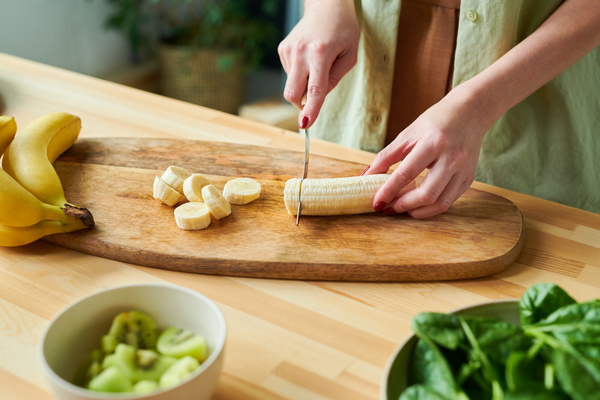 A woman making a smoothie cuts a banana into slices with a small knife on a wooden cutting board next to which are other ingredients for the drink