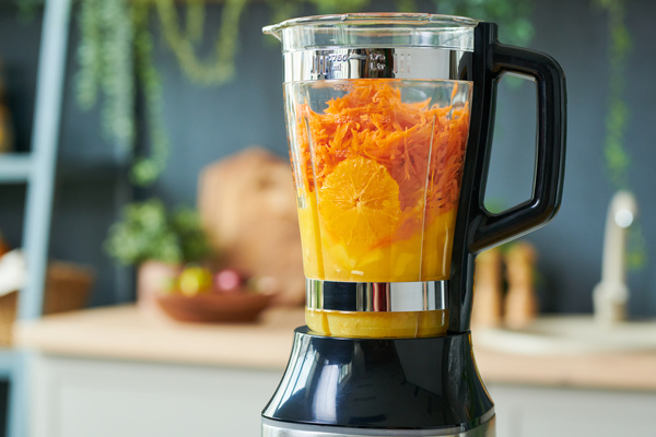There is a peeled and sliced orange and grated carrots for making smoothie in a jug of a blender with a black handle