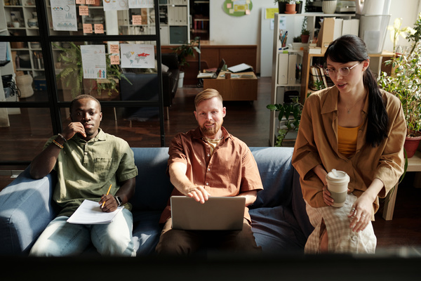 A team of startuppers consisting of two men in shirts one of whom is holding a pencil and documents and the second is holding a laptop and a woman with dark hair with a cup of coffee sitting on the couch discussing the project