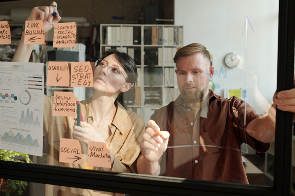 Partners in a startup project standing at the glass board with stickers in a light coworking with felt-tip pens make notes related to work