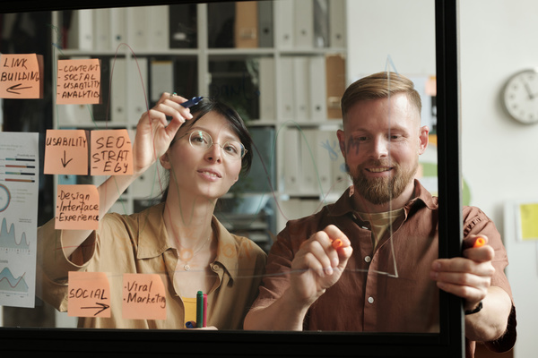 Colleagues on a startup project standing at the glass board with stickers in a bright office draw work-related graphs with felt-tip pens