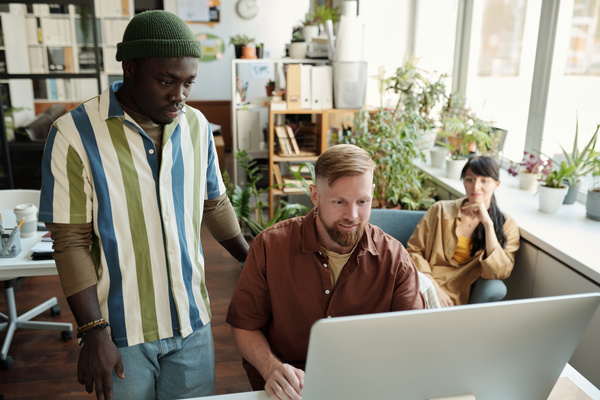 A man with blond hair and beard is working on a computer sitting at a desk in the office and his working partner in a striped shirt is standing next to him a woman with dark hair is sitting behind them