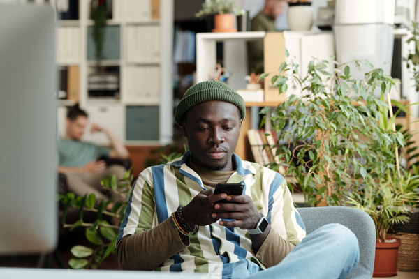 A guy in a dark green hat and striped shirt is sitting on a chair in a light coworking in a surfing the Internet on a black smartphone