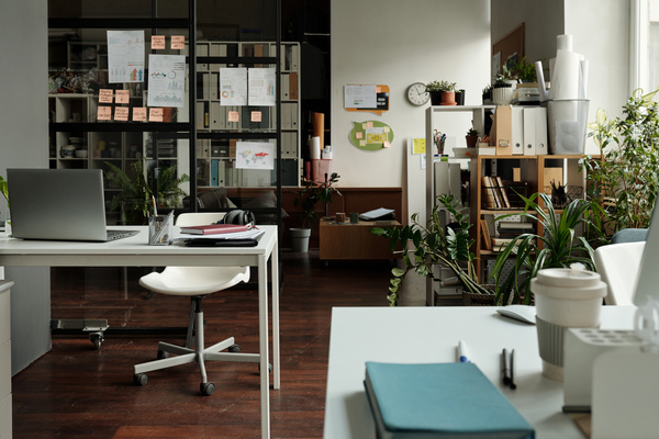 The interior of a bright startup coworking with a white desk in the foreground on which there are documents there is a cup and a silver laptop and stationery in the organizer