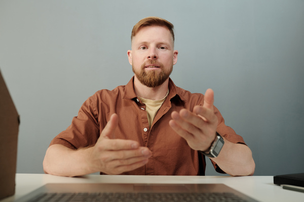 A young blond man in a shirt communicates with colleagues on work issues via a video call via a laptop actively gesticulating