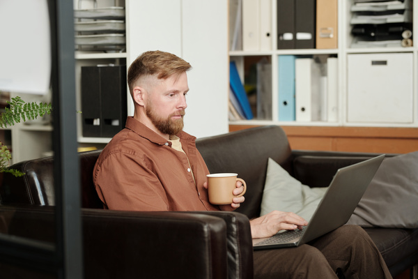 A male startupper with blond hair and beard is sitting on a dark sofa with a mug in his hand and typing on a laptop that is on his lap