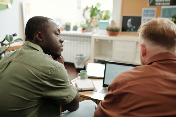 Two young men are sitting in a light coworking in shirts in front of a laptop standing on a table and talking