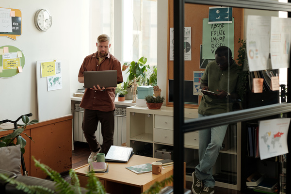 Male colleagues in green and brown shirts in a light coworking with plants standing work on a laptop and on a tablet