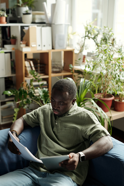 A man with black afro hair and beard dressed in a swamp-colored shirt sitting on a blue sofa with a tablet and documents in his hands looks at them