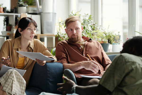 A woman with black hair gathered in a ponytail holding documents and a pen in her hands talks to a working partner sitting opposite her and a young man with a beard who listens to their conversation
