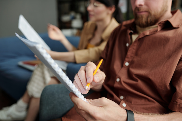 A man with a beard dressed in a brick-colored shirt holding documents held together by a spring makes notes in them sitting next to a woman who is also studying work papers