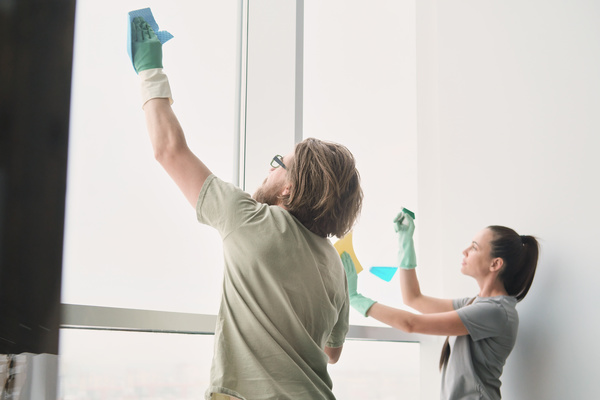 A husband with glasses and his wife with long hair gathered in a ponytail in home clothes and rubber gloves wiping a large window with household chemicals and cleaning cloths