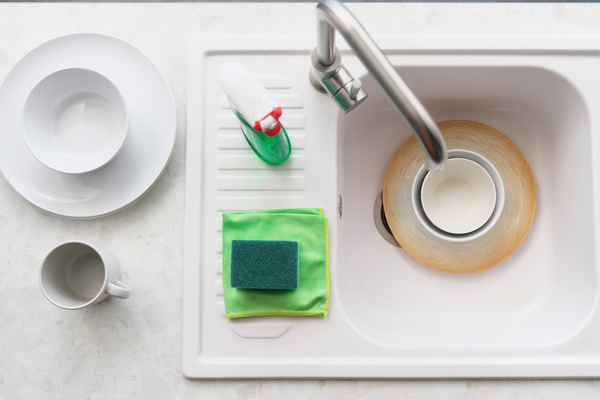 Plates in a white sink with detergent in a spray bottle and a green sponge and rag on it
