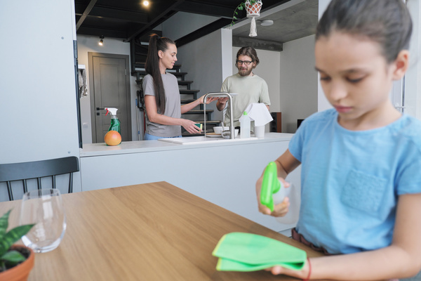 A woman with long hair in a ponytail standing at the sink passes a washed plate to her husband with a kitchen towel and their daughter applies detergent on a rag to wipe the table