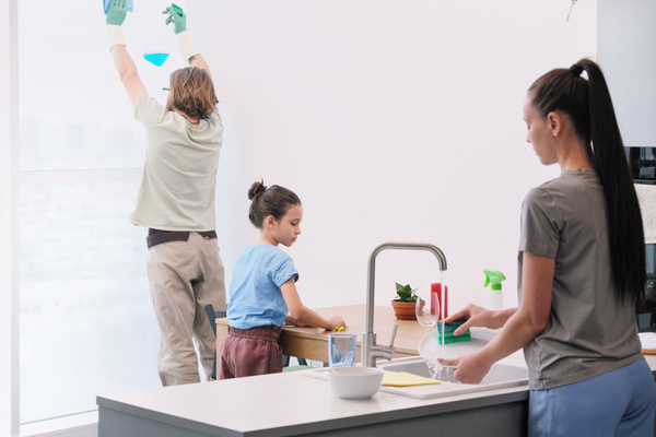 A woman with gathered hair washing a plate with a green sponge in the kitchen with daughter wiping the table and husband washing the window