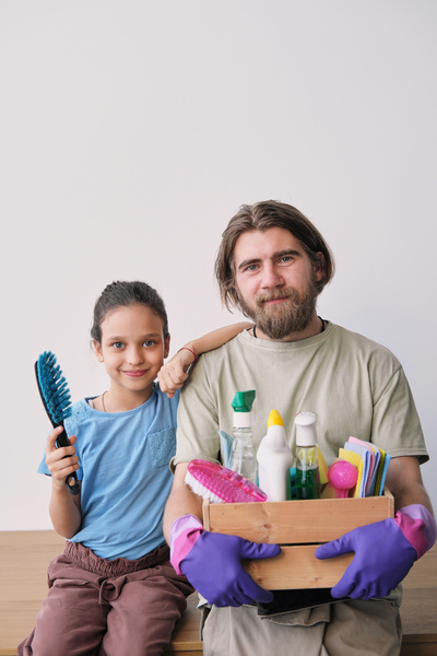 A girl with a cleaning brush is sitting next to her father with a beard and purple rubber gloves holding a cleaning set in a wooden box
