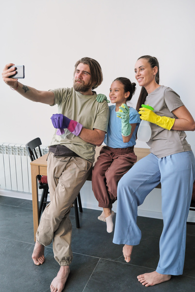 A man in purple rubber gloves takes a selfie with his wife and daughter with a cleaning kit sitting on a table