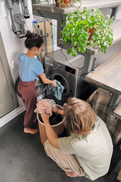 The father squatting gives daughter clothes from the laundry basket and she puts it in the washer