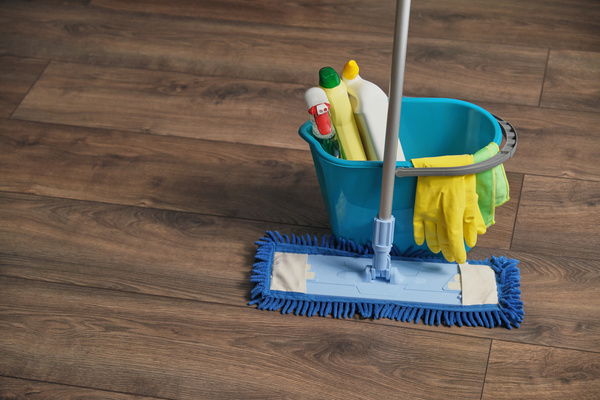 A mop with a microfiber nozzle is placed next to a bright blue bucket with detergents and yellow rubber gloves on a dark parquet floor