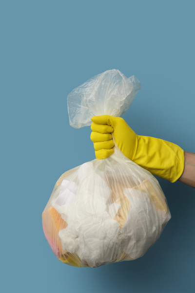 A white cellophane garbage bag filled with garbage is held on a blue background in the hand in a yellow rubber glove