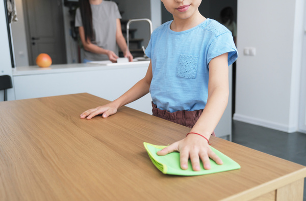 A girl in a blue blouse wipes a wooden kitchen table with a green microfiber cloth