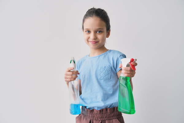 A girl in a blue blouse with her hair gathered smiling holds blue and green detergents in sprayers