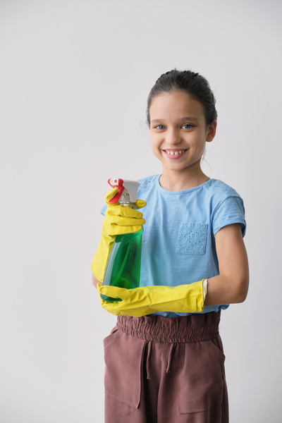 A girl with yellow rubber gloves on her hands smiling holds a green cleaning agent in a spray bottle