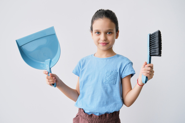 A girl with her hair in a bun smiling holds a blue dustpan and a floor cleaning brush with black bristles