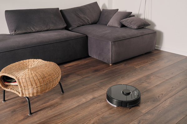 Black robot vacuum cleaner hoovers parquet in the living room with a gray corner sofa