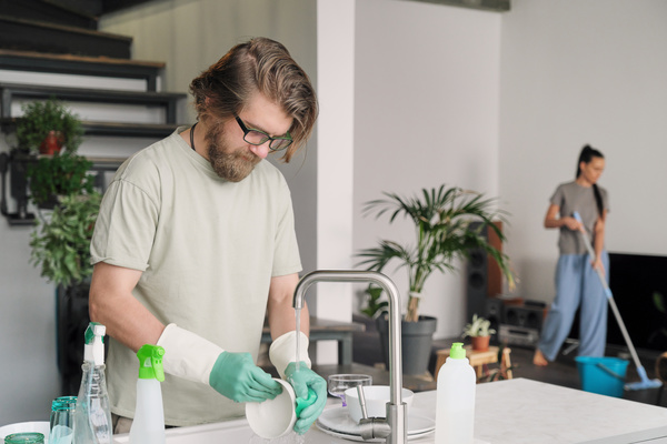 A man with a beard in beige clothes and green rubber gloves washes a white bowl with a sponge with detergent