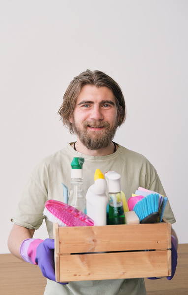 A man with a beard smiling holds a wooden box with household chemicals and cleaning equipment