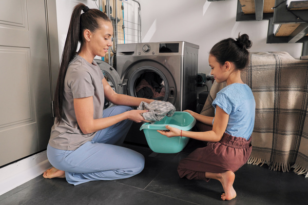 A woman with long hair in a ponytail puts clothes from a washing machine into a turquoise basin held by her daughter