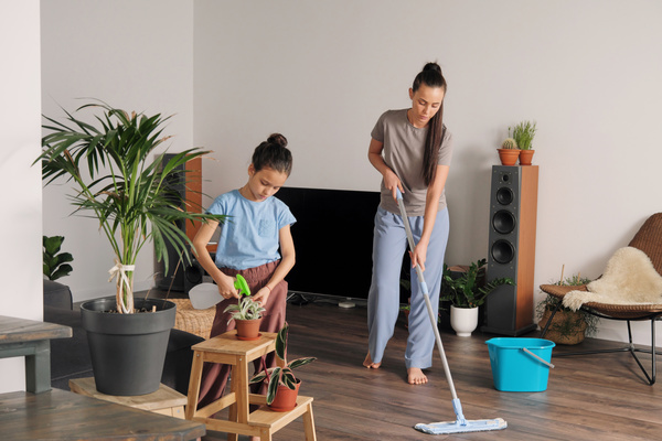A woman with long hair gathered in a ponytail washes the floor with a mop and her daughter sprays water on domestic plants