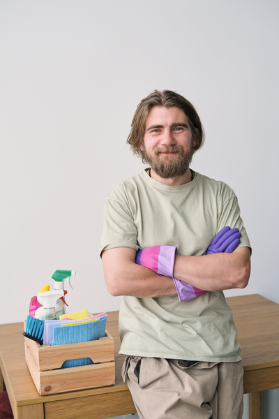 A man with a beard wearing beige shades and purple rubber gloves stands with his arms crossed on his chest next to a table with a cleaning kit in a wooden box on it