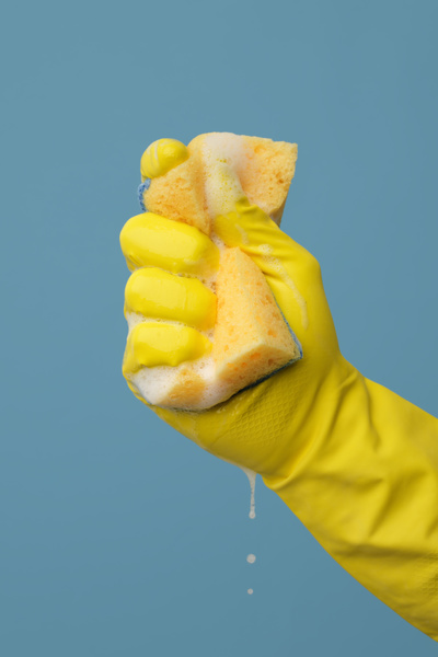 Yellow foam sponge for washing dishes in a clenched hand in a yellow rubber glove on a blue background
