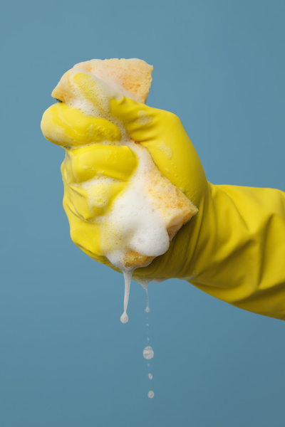 A yellow foam sponge for washing dishes is squeezed in the hand in a yellow rubber glove on a blue background