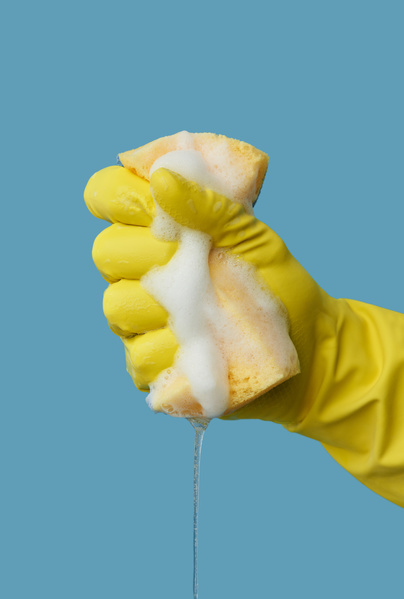 A yellow soap sponge for cleaning is squeezed out in a hand in a yellow rubber glove on a blue background
