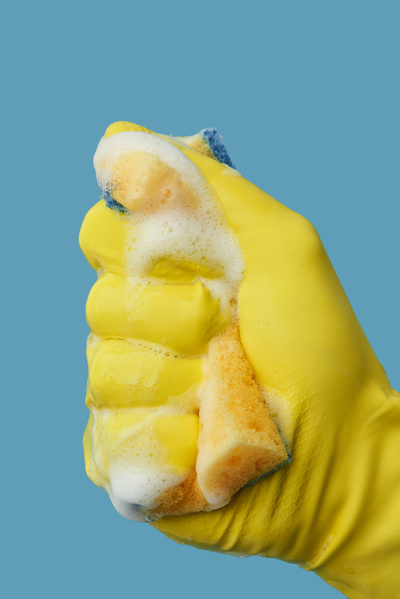 A hand in a yellow rubber glove clutching a foam sponge for washing dishes on a blue background