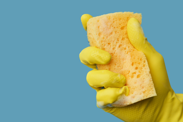 A hand in a yellow rubber glove holding a soap sponge for washing dishes on a blue background
