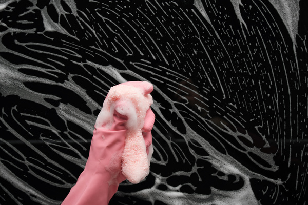A hand in a pink rubber glove clutching a foamy terry sponge for washing dishes on a dark background with foam stains