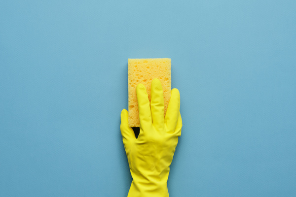 A yellow cleaning sponge is held with a hand in a yellow rubber glove on a blue background