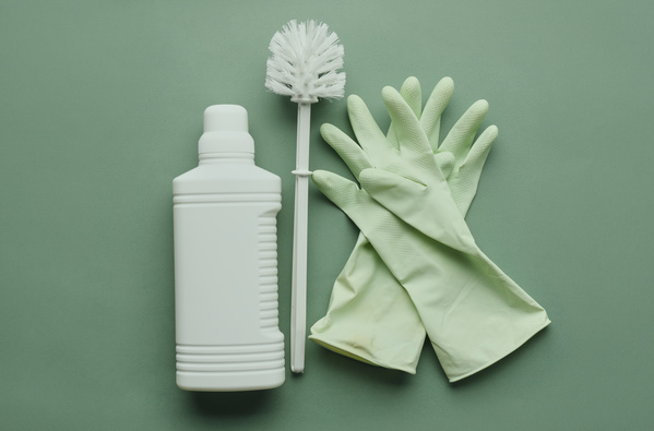 A white bottle with household chemicals a toilet brush and green rubber gloves on a green surface