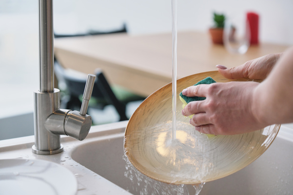 A flat plate of light color is washed with a yellow sponge in the kitchen under a stream of water from a metal faucet