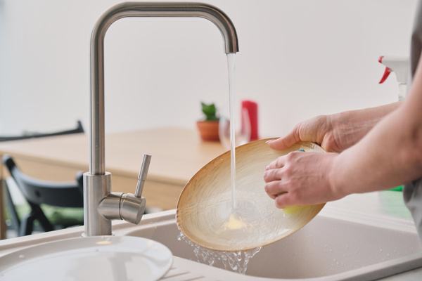 A flat round platter is washed with a sponge under a stream of water from a kitchen metal faucet