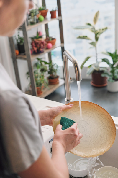 A flat round dish is cleaned with a sponge under a stream of water from a kitchen metal faucet