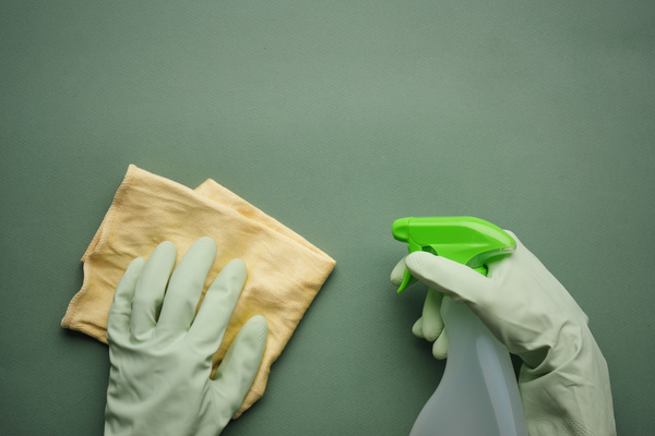 A yellow microfiber cloth and window cleaner are held with hands in green rubber gloves on a green background